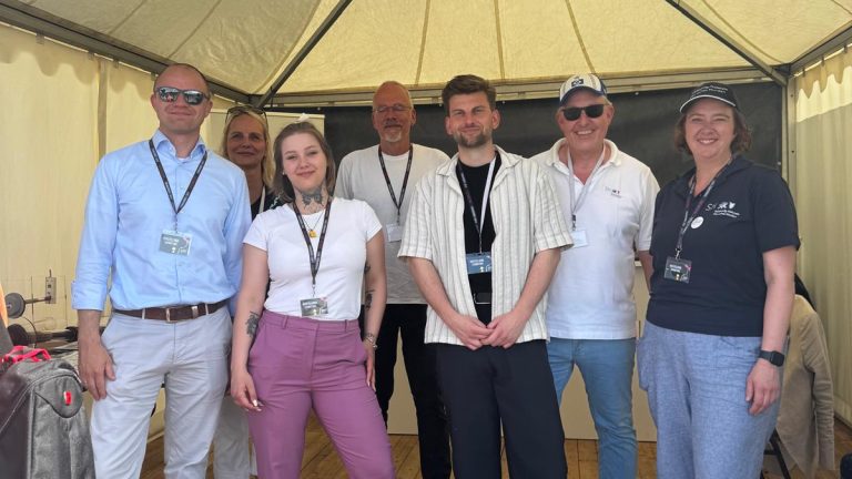 Eine Gruppe von sieben Personen steht unter einem weißen Pavillon mit der Aufschrift „BKZ.SH, ZVO & LAND SH“. Sie lächeln und tragen Namensschilder. Im Vordergrund sind Holzboden und ein Tisch mit Broschüren zu sehen.