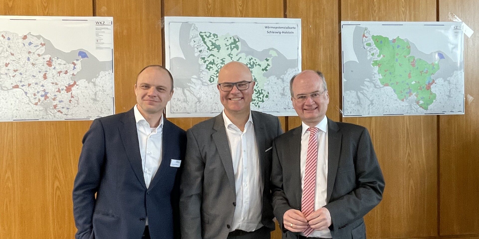 IMG 0447 Drei Männer in Anzügen stehen in einem Konferenzraum vor holzverkleideten Wänden. An der Wand hängen Karten. Im Vordergrund sind schwarze Bürostühle und ein Tisch mit Dokumenten sichtbar. Ein Projektor ist an der Decke montiert.