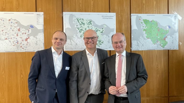 Drei Männer in Anzügen stehen in einem Konferenzraum vor holzverkleideten Wänden. An der Wand hängen Karten. Im Vordergrund sind schwarze Bürostühle und ein Tisch mit Dokumenten sichtbar. Ein Projektor ist an der Decke montiert.