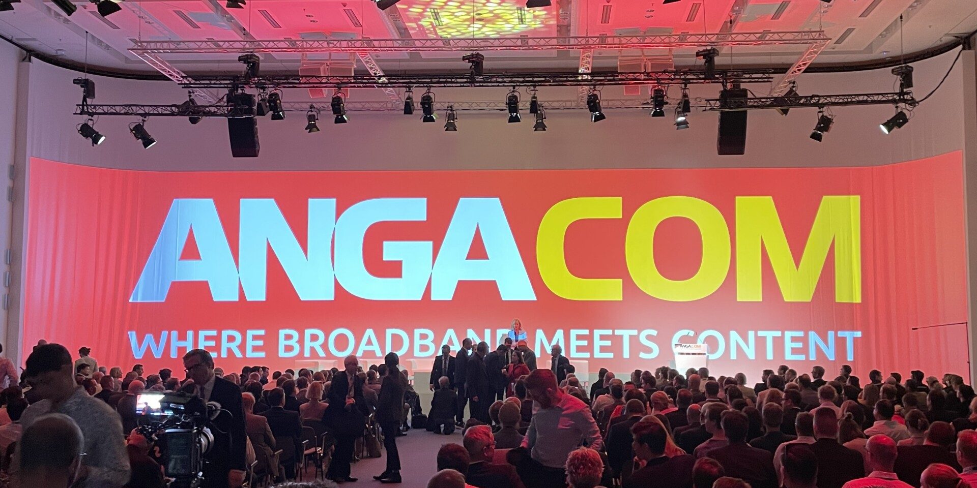 Eine große Konferenzhalle mit vielen sitzenden Personen. An der Vorderseite ist eine riesige Projektionswand zu sehen, die mit "ANGACOM" in großen Buchstaben und dem Slogan "Where Broadband Meets Content" beleuchtet ist.