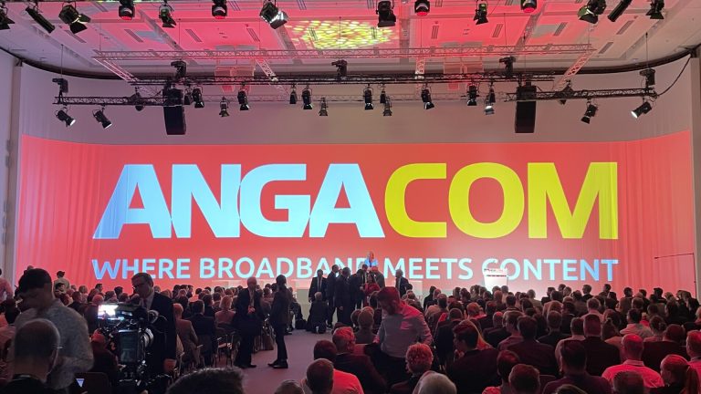 Eine große Konferenzhalle mit vielen sitzenden Personen. An der Vorderseite ist eine riesige Projektionswand zu sehen, die mit "ANGACOM" in großen Buchstaben und dem Slogan "Where Broadband Meets Content" beleuchtet ist.