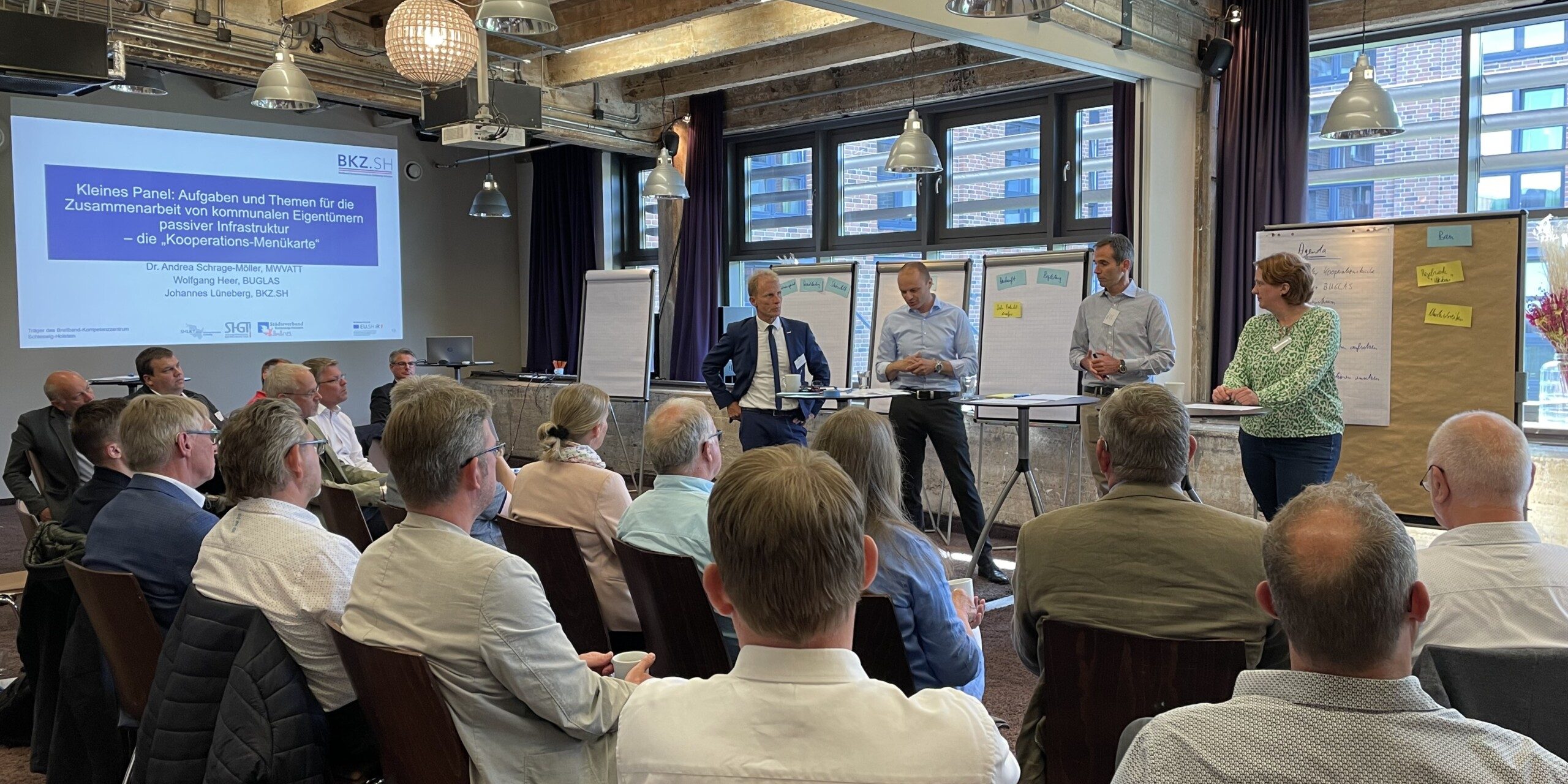 Ein Gruppenraum mit Menschen, die einer Präsentation folgen. Vorne stehen fünf Personen an Flipcharts und einem Bildschirm. Die Teilnehmer sitzen in Reihen und schauen nach vorn. Raum mit Holzdecke und großen Fenstern.