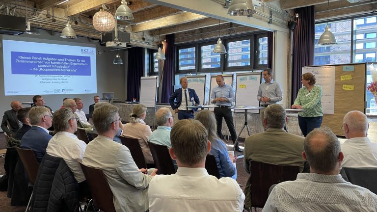 Ein Gruppenraum mit Menschen, die einer Präsentation folgen. Vorne stehen fünf Personen an Flipcharts und einem Bildschirm. Die Teilnehmer sitzen in Reihen und schauen nach vorn. Raum mit Holzdecke und großen Fenstern.