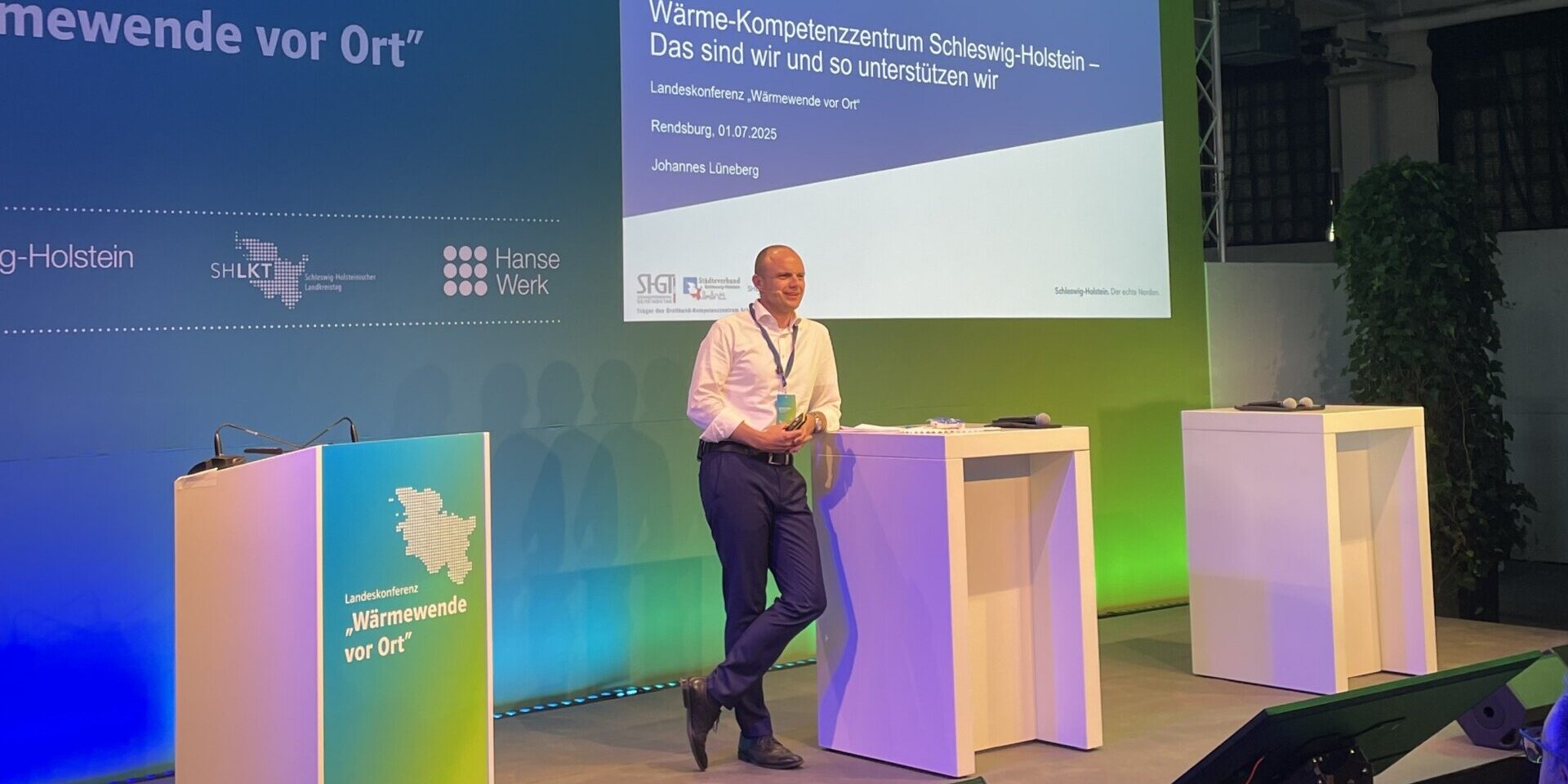 Ein Mann steht auf einer Bühne und spricht vor Publikum. Im Hintergrund ist eine Leinwand mit dem Logo der BKZ.SH und einem Vortragstitel zu "Wärmewende vor Ort" in Schleswig-Holstein. Die Beleuchtung ist in Blau- und Grüntönen gehalten.