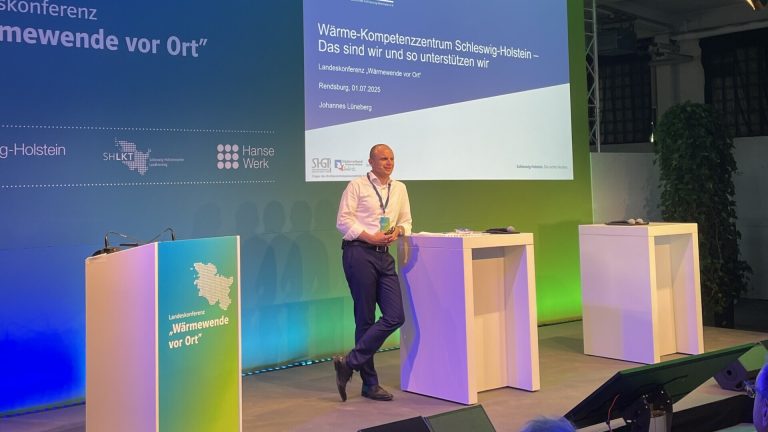 Ein Mann steht auf einer Bühne und spricht vor Publikum. Im Hintergrund ist eine Leinwand mit dem Logo der BKZ.SH und einem Vortragstitel zu "Wärmewende vor Ort" in Schleswig-Holstein. Die Beleuchtung ist in Blau- und Grüntönen gehalten.