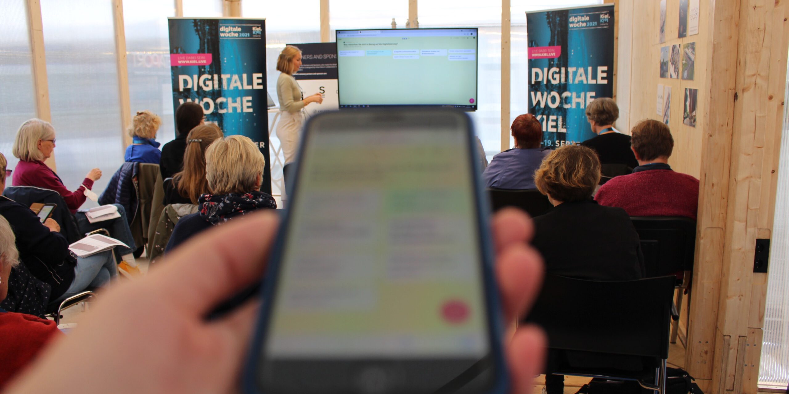 Eine Frau präsentiert vor einer Gruppe in einem Raum mit Glaswänden. Im Vordergrund hält jemand ein Smartphone in der Hand, das unscharf ist. Stühle sind besetzt, und zwei Roll-Ups mit der Aufschrift "Digitale Woche" sind sichtbar.