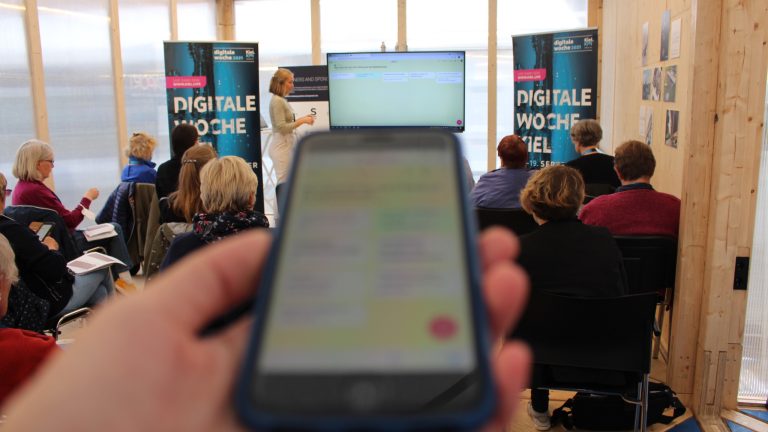 Eine Frau präsentiert vor einer Gruppe in einem Raum mit Glaswänden. Im Vordergrund hält jemand ein Smartphone in der Hand, das unscharf ist. Stühle sind besetzt, und zwei Roll-Ups mit der Aufschrift "Digitale Woche" sind sichtbar.