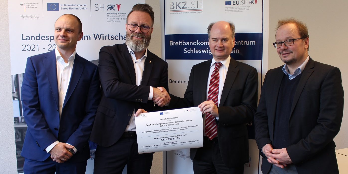 Vier Männer stehen vor Roll-Up Bannern. Zwei der Männer in der Mitte schütteln sich lächelnd die Hände und halten zusammen ein großes Zertifikat. Alle tragen Anzüge.