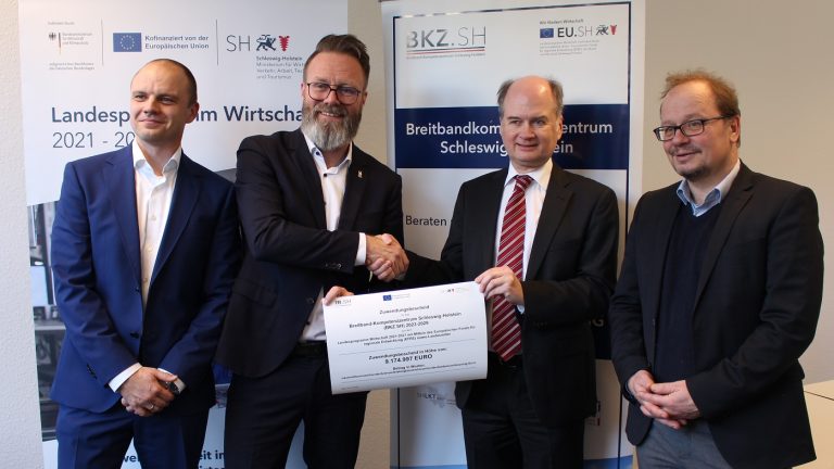 Vier Männer stehen vor Roll-Up Bannern. Zwei der Männer in der Mitte schütteln sich lächelnd die Hände und halten zusammen ein großes Zertifikat. Alle tragen Anzüge.