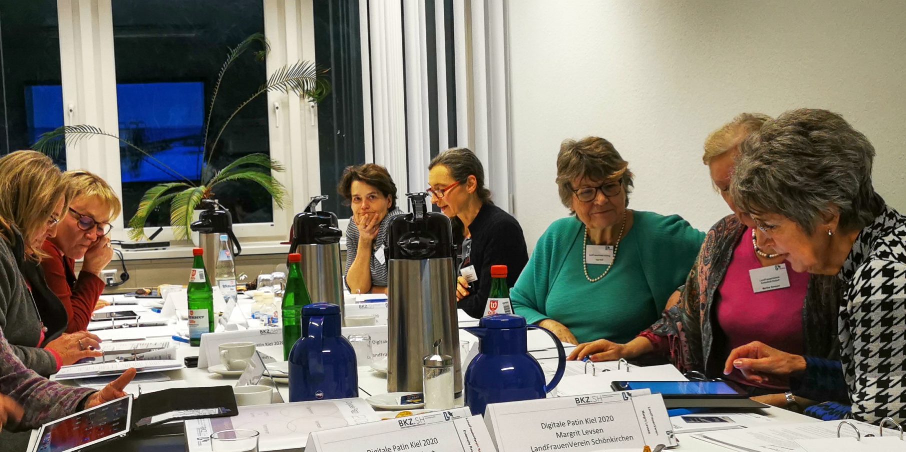 IMG 20200205 171409 Bearbeitet Eine Gruppe von zehn Personen sitzt an einem Konferenztisch in einem Raum. Sie arbeiten an Laptops und Papieren. Auf dem Tisch stehen Thermoskannen, Flaschen und Namensschilder. Einige Personen lächeln, der Raum hat Fenster im Hintergrund.