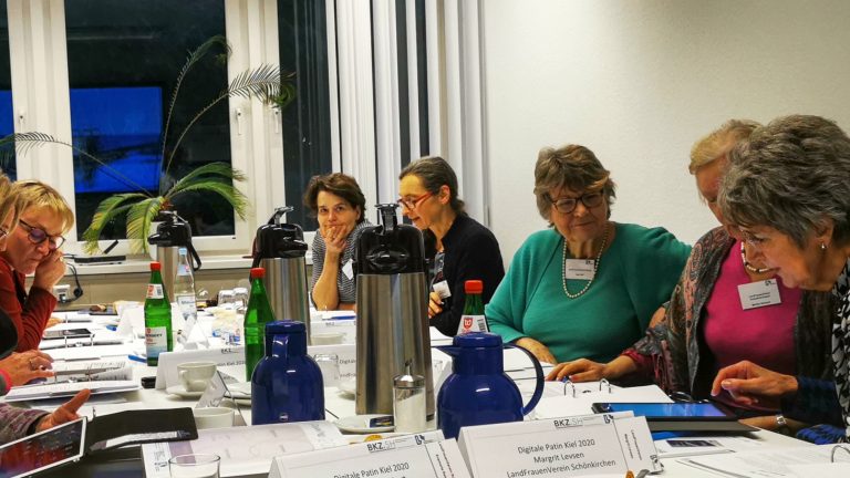 Eine Gruppe von zehn Personen sitzt an einem Konferenztisch in einem Raum. Sie arbeiten an Laptops und Papieren. Auf dem Tisch stehen Thermoskannen, Flaschen und Namensschilder. Einige Personen lächeln, der Raum hat Fenster im Hintergrund.