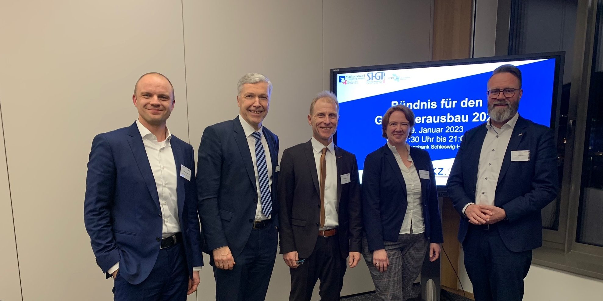 Image Kopie Fünf Personen stehen lächelnd in einem Raum. Im Hintergrund ist ein Bildschirm mit der Aufschrift "Bündnis für den Glasfaserausbau 2023" zu sehen. Die Personen tragen Anzüge und stehen nebeneinander vor einem Fenster.