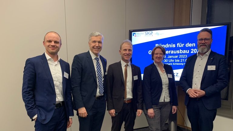 Fünf Personen stehen lächelnd in einem Raum. Im Hintergrund ist ein Bildschirm mit der Aufschrift "Bündnis für den Glasfaserausbau 2023" zu sehen. Die Personen tragen Anzüge und stehen nebeneinander vor einem Fenster.