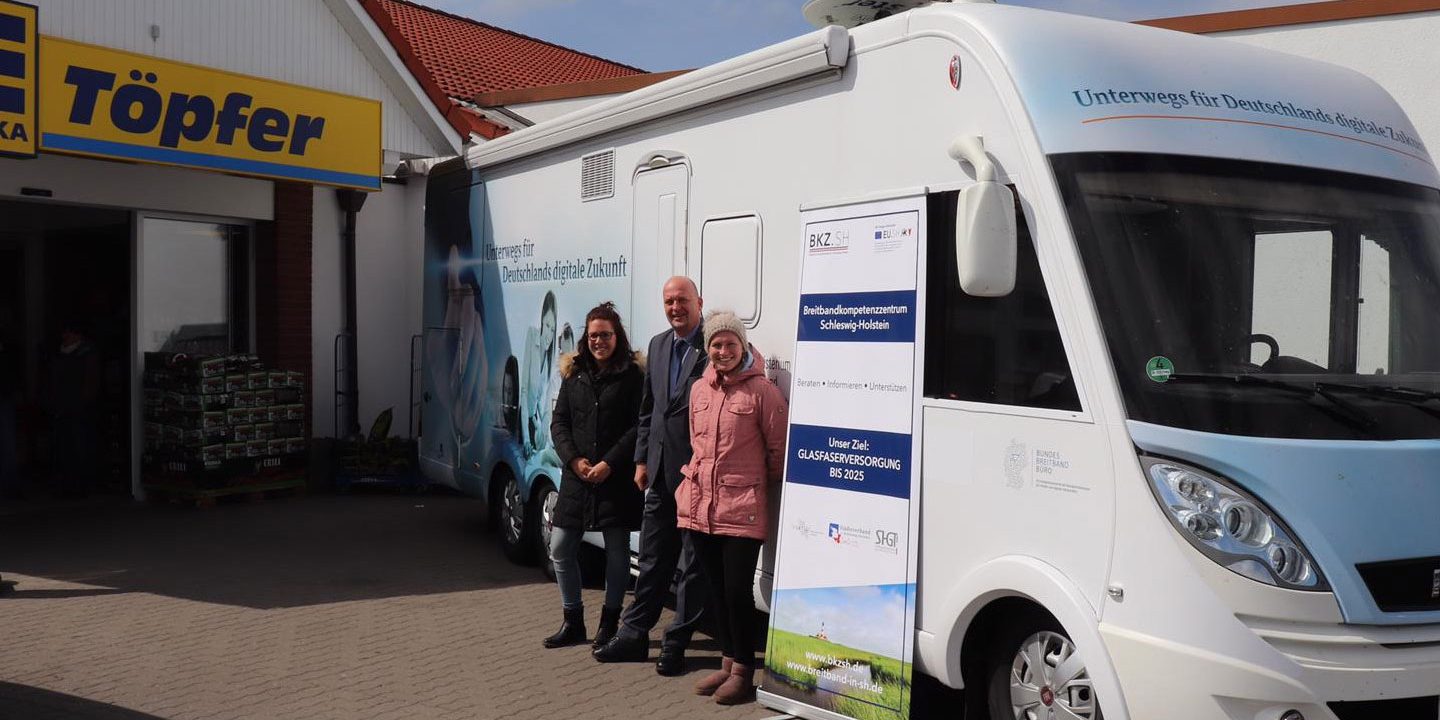 Eine Gruppe von drei Menschen steht vor einem weißen Infomobil mit Aufklebern. Neben dem Fahrzeug ist ein rollbares Werbebanner. Im Hintergrund ist ein Gebäude mit der Aufschrift "Töpfer" zu sehen.