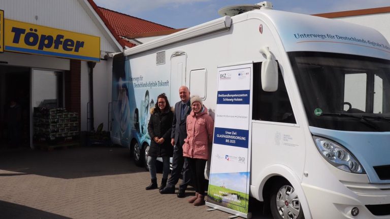 Eine Gruppe von drei Menschen steht vor einem weißen Infomobil mit Aufklebern. Neben dem Fahrzeug ist ein rollbares Werbebanner. Im Hintergrund ist ein Gebäude mit der Aufschrift "Töpfer" zu sehen.