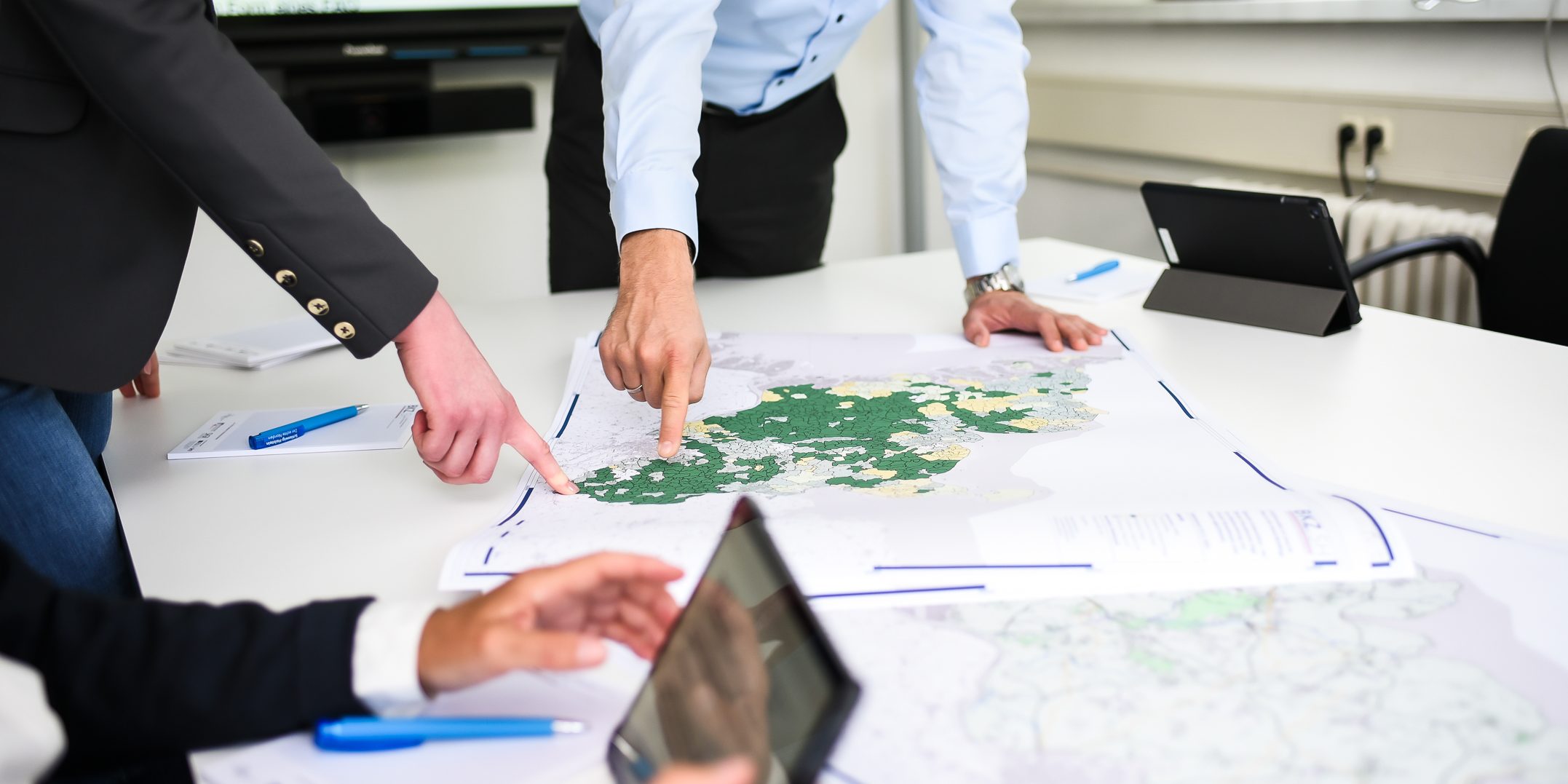 KT7 0072 Personen stehen um einen Tisch und zeigen auf eine große Landkarte. Auf dem Tisch liegen Stifte und Dokumente. Im Hintergrund sind ein Bildschirm und Fenster zu sehen.