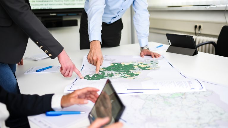 Personen stehen um einen Tisch und zeigen auf eine große Landkarte. Auf dem Tisch liegen Stifte und Dokumente. Im Hintergrund sind ein Bildschirm und Fenster zu sehen.