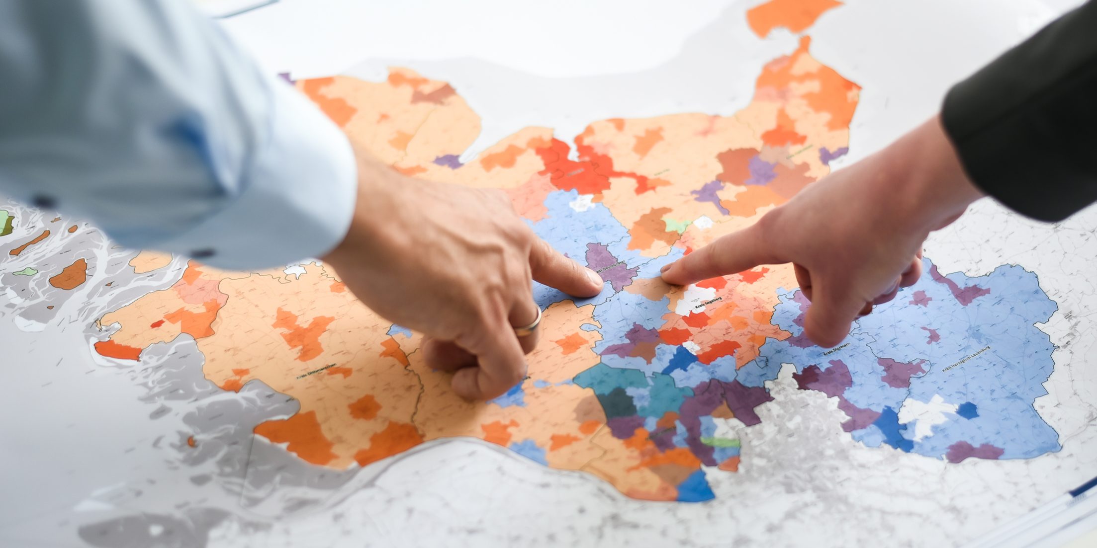 KT7 0113 Zwei Hände zeigen auf eine bunte Karte mit unterschiedlichen Regionen in Orange-, Blau- und Rottönen. Die Karte liegt auf einem Tisch, und die Hände scheinen Details zu diskutieren oder zu analysieren.