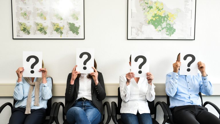 Vier Personen sitzen auf Stühlen in einem Raum und halten Schilder mit großen Fragezeichen vor ihren Gesichtern. Hinter ihnen sind zwei Landkarten an der Wand aufgehängt. Alle tragen formelle Kleidung.