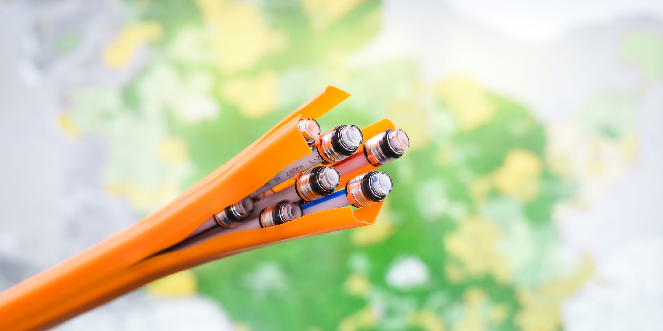 KT7 0416 Das Bild zeigt ein Bündel von orangefarbenen Kabeln, deren Enden freiliegen und mehrere silberne Steckverbindungen sichtbar sind. Der Hintergrund ist unscharf mit grünen und gelben Farbtönen.