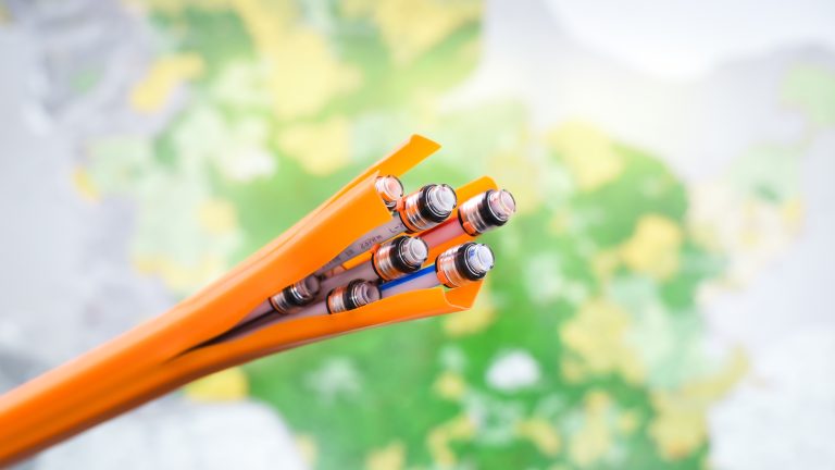 Das Bild zeigt ein Bündel von orangefarbenen Kabeln, deren Enden freiliegen und mehrere silberne Steckverbindungen sichtbar sind. Der Hintergrund ist unscharf mit grünen und gelben Farbtönen.