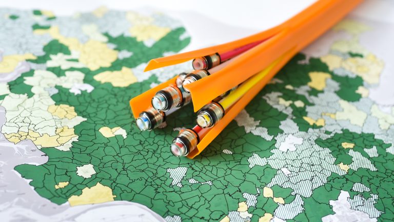 Mehrere orangefarbene, geöffnete Rohre mit bunten Kabeln darin liegen auf einer Karte. Die Karte zeigt unterschiedliche Regionen in Grün- und Gelbtönen, die an politische oder geografische Grenzen erinnern.