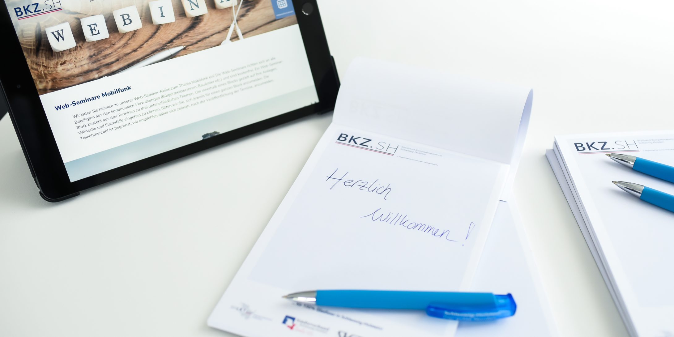 KT7 0522 Ein Tablet zeigt eine Webseite mit dem Wort "Webinar". Daneben liegen Schreibblöcke mit der Aufschrift "Herzlich Willkommen!" und Kugelschreiber auf einem weißen Tisch.