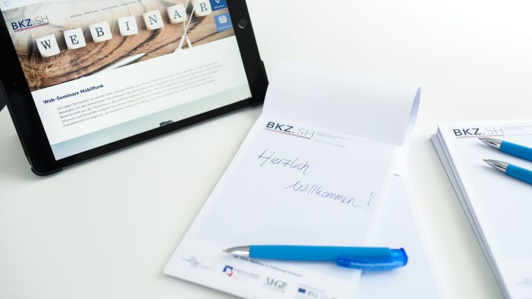 Ein Tablet zeigt eine Webseite mit dem Wort "Webinar". Daneben liegen Schreibblöcke mit der Aufschrift "Herzlich Willkommen!" und Kugelschreiber auf einem weißen Tisch.