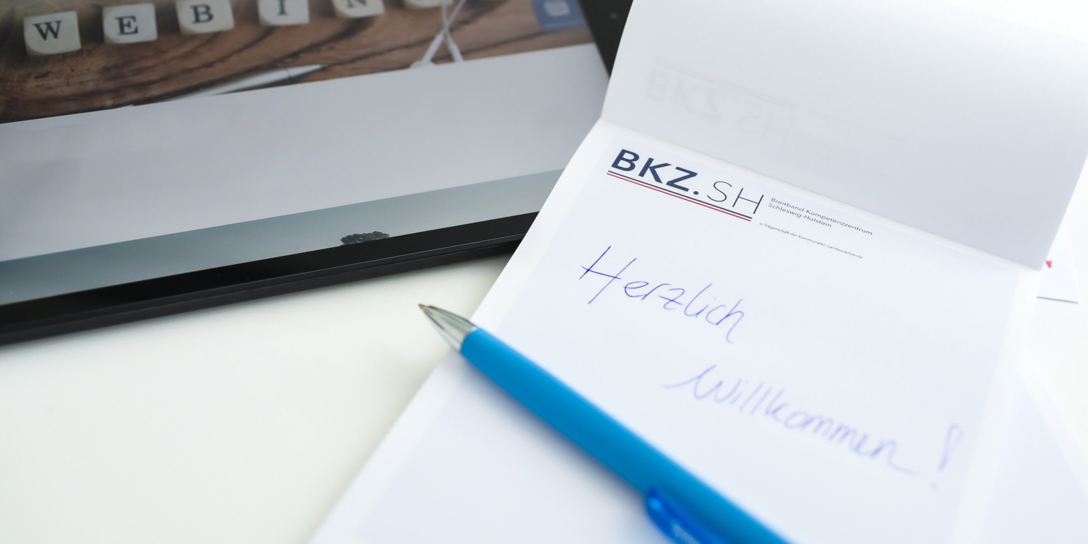 Ein Tablet mit der Anzeige "WEBINAR" liegt neben einem Notizblock mit dem Logo "BKZ.SH". Auf dem Block steht in blauer Schrift "Herzlich Willkommen!". Ein blauer Kugelschreiber liegt darauf.