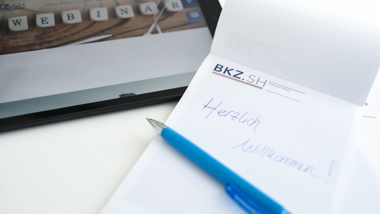 Ein Tablet mit der Anzeige "WEBINAR" liegt neben einem Notizblock mit dem Logo "BKZ.SH". Auf dem Block steht in blauer Schrift "Herzlich Willkommen!". Ein blauer Kugelschreiber liegt darauf.