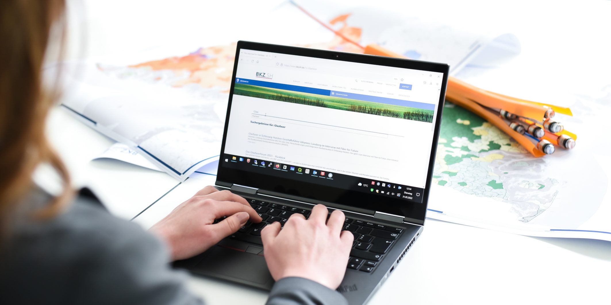 Eine Person tippt auf einem Laptop, der auf einem Tisch steht. Im Hintergrund liegen geöffnete Atlanten und orangefarbene Bleistifte. Auf dem Laptop ist eine Website mit Text und Bildern geöffnet.