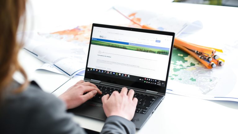 Eine Person tippt auf einem Laptop, der auf einem Tisch steht. Im Hintergrund liegen geöffnete Atlanten und orangefarbene Bleistifte. Auf dem Laptop ist eine Website mit Text und Bildern geöffnet.