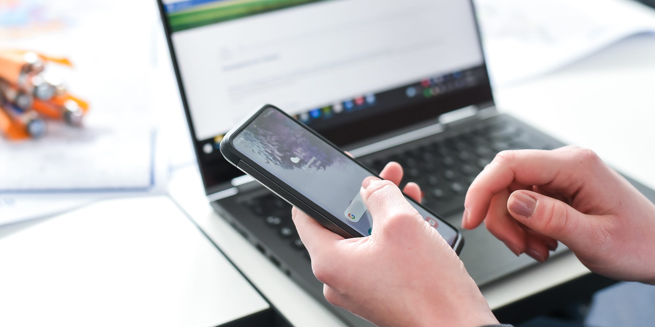 Eine Person hält ein Smartphone in der Hand und blickt darauf, während ein Laptop mit geöffneter Webseite im Hintergrund auf einem Tisch steht. Die Umgebung ist hell und modern gestaltet.