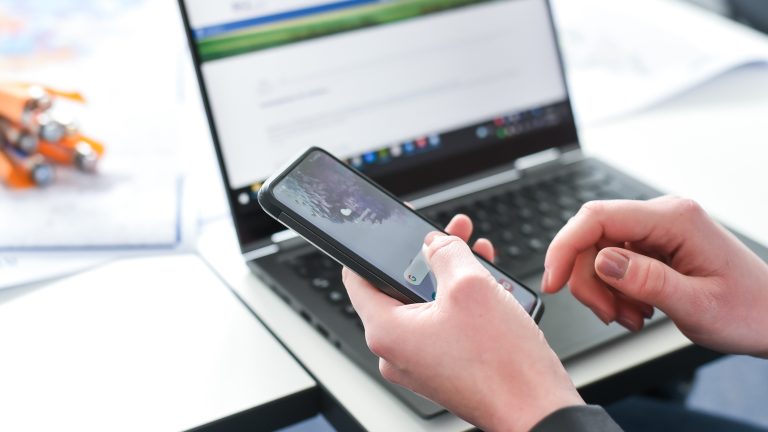 Eine Person hält ein Smartphone in der Hand und blickt darauf, während ein Laptop mit geöffneter Webseite im Hintergrund auf einem Tisch steht. Die Umgebung ist hell und modern gestaltet.