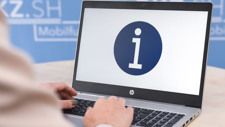 Eine Person sitzt vor einem Laptop, auf dessen Bildschirm ein blaues Informationssymbol mit einem weißen "i" angezeigt wird. Der Hintergrund ist blau mit unscharfen weißen Texten.