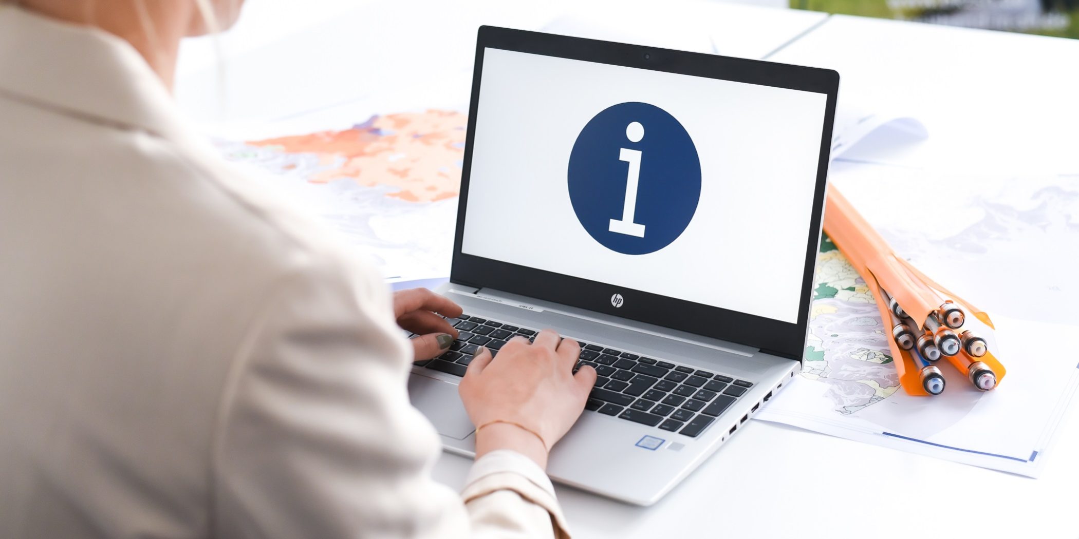 Info Eine Person sitzt an einem Schreibtisch und tippt auf einem Laptop. Auf dem Bildschirm ist ein blaues Kreis-Symbol mit einem weißen „i“ zu sehen. Neben dem Laptop liegen gerollte Pläne oder Landkarten.