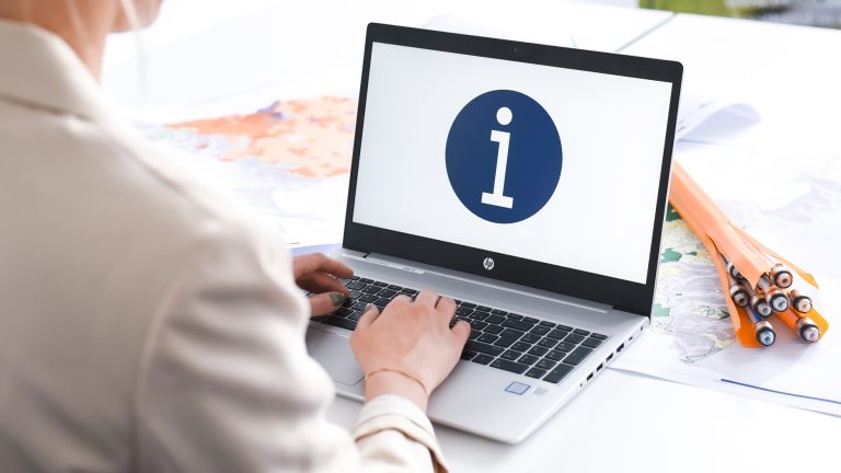 Eine Person sitzt an einem Schreibtisch und tippt auf einem Laptop. Auf dem Bildschirm ist ein blaues Kreis-Symbol mit einem weißen „i“ zu sehen. Neben dem Laptop liegen gerollte Pläne oder Landkarten.