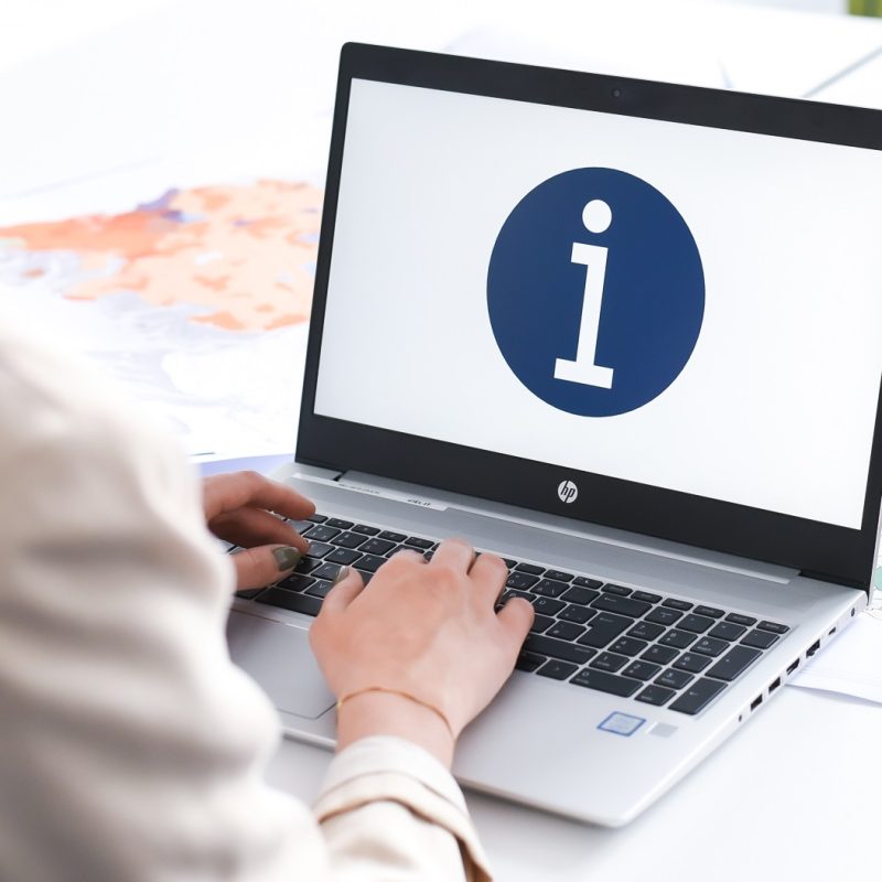 Eine Person sitzt an einem Schreibtisch und tippt auf einem Laptop. Auf dem Bildschirm ist ein blaues Kreis-Symbol mit einem weißen „i“ zu sehen. Neben dem Laptop liegen gerollte Pläne oder Landkarten.