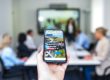 Eine Hand hält ein Smartphone, das eine Webseite zeigt. Im Hintergrund sitzen Menschen an einem Konferenztisch vor einem großen Bildschirm, der eine Präsentation darstellt. Die Szene ist hell erleuchtet.