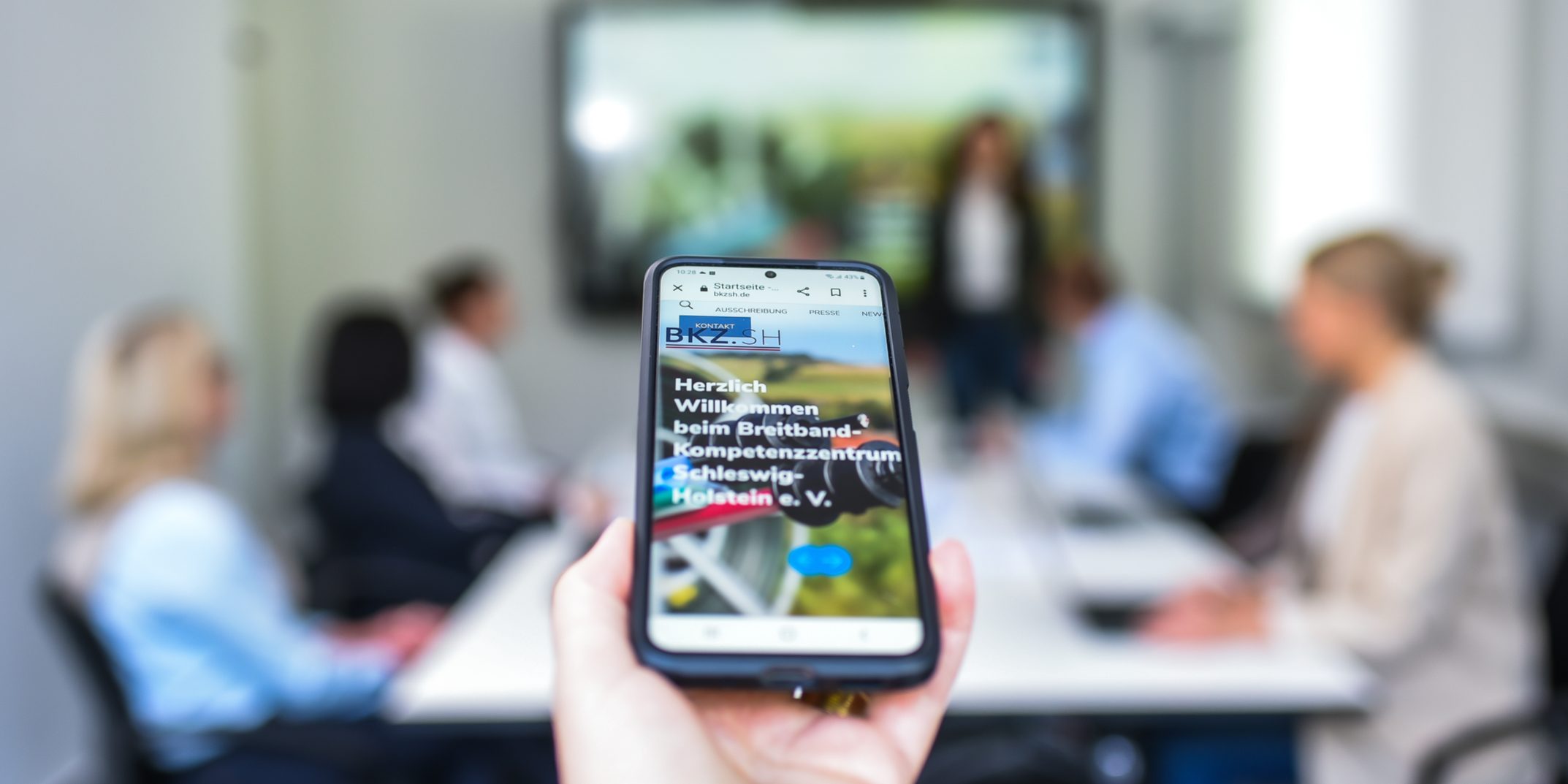 KT7 9985 Eine Hand hält ein Smartphone, das eine Webseite zeigt. Im Hintergrund sitzen Menschen an einem Konferenztisch vor einem großen Bildschirm, der eine Präsentation darstellt. Die Szene ist hell erleuchtet.