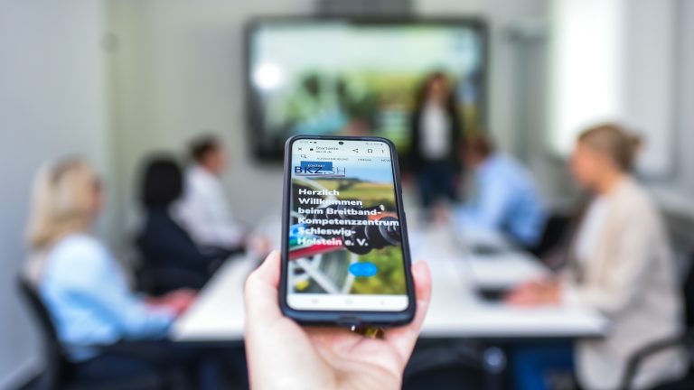 Eine Hand hält ein Smartphone, das eine Webseite zeigt. Im Hintergrund sitzen Menschen an einem Konferenztisch vor einem großen Bildschirm, der eine Präsentation darstellt. Die Szene ist hell erleuchtet.