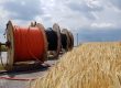 Drei große Kabeltrommeln stehen nebeneinander auf einer Asphaltfläche. Zwei Trommeln haben schwarze Kabel, eine hat orange Kabel. Im Vordergrund ist ein Weizenfeld, darüber ein bewölkter Himmel.