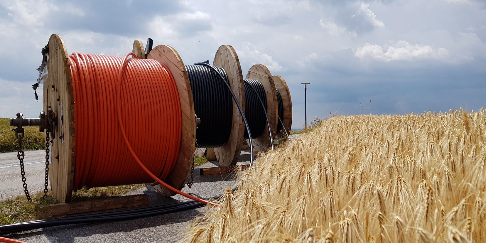 Drei große Kabeltrommeln stehen nebeneinander auf einer Asphaltfläche. Zwei Trommeln haben schwarze Kabel, eine hat orange Kabel. Im Vordergrund ist ein Weizenfeld, darüber ein bewölkter Himmel.