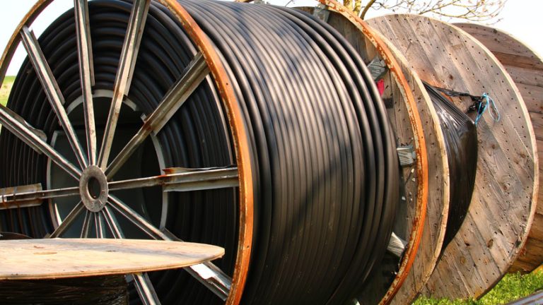 Große Holztrommeln mit aufgerollten schwarzen Kabeln liegen auf einer grünen Wiese. Im Hintergrund ist ein kahler Baum zu sehen.
