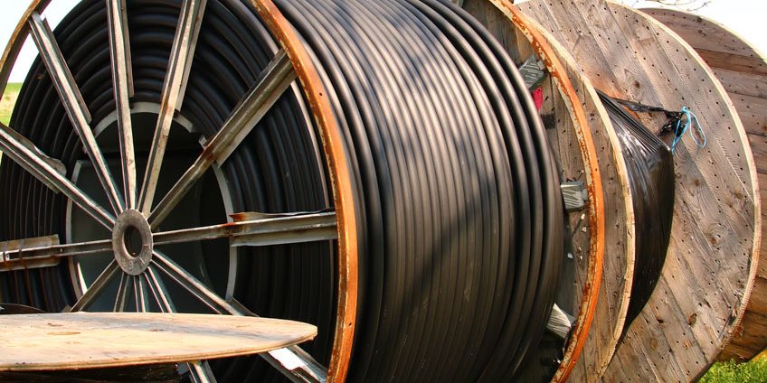 Kabeltrommeln Schwarz MainImage Große Holztrommeln mit aufgerollten schwarzen Kabeln liegen auf einer grünen Wiese. Im Hintergrund ist ein kahler Baum zu sehen.