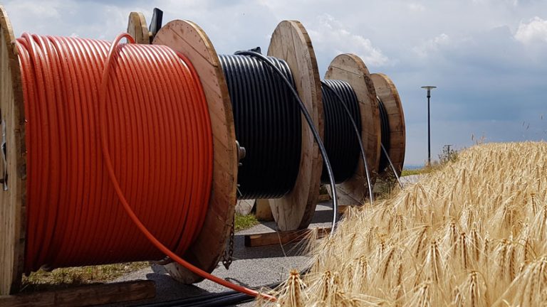 Große Holztrommeln mit aufgerollten Kabeln in Orange und Schwarz stehen unter bewölktem Himmel auf einer Straße neben einem goldenen Getreidefeld. Ein Kabel verläuft über die Straße ins Feld.