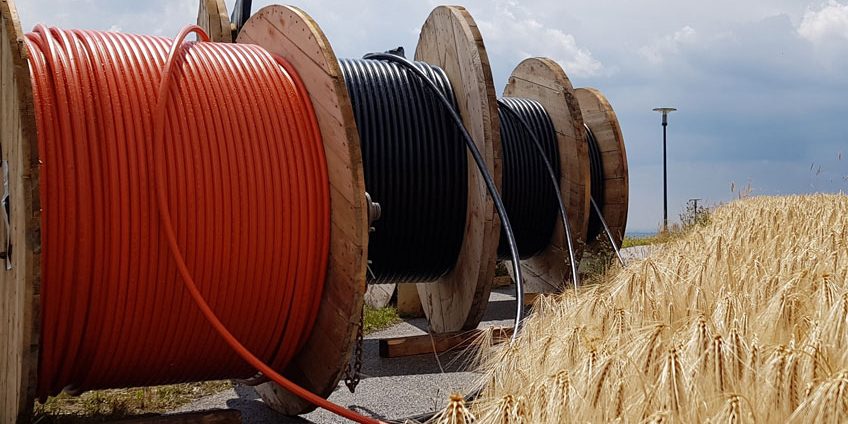 Große Holztrommeln mit aufgerollten Kabeln in Orange und Schwarz stehen unter bewölktem Himmel auf einer Straße neben einem goldenen Getreidefeld. Ein Kabel verläuft über die Straße ins Feld.