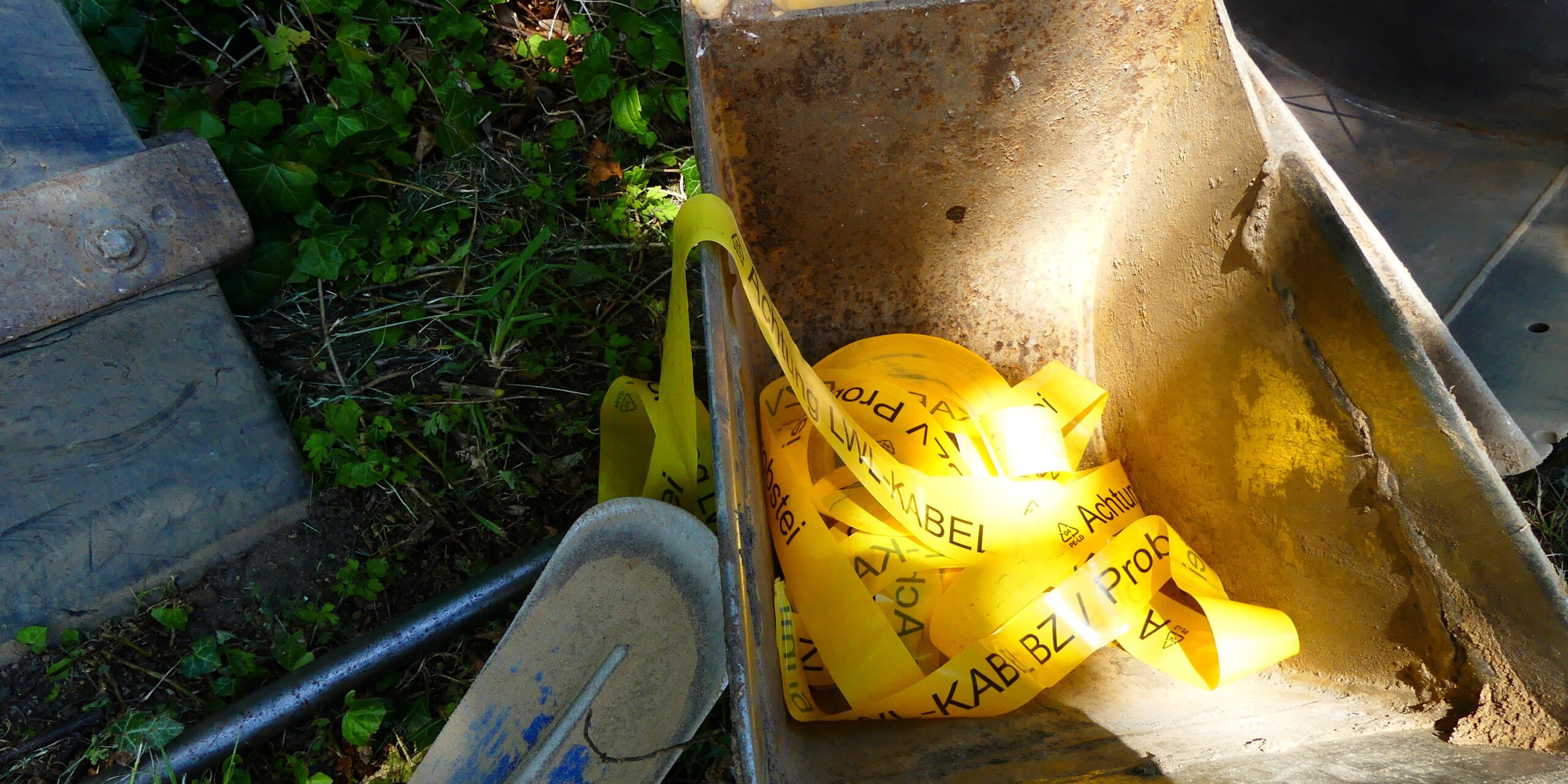 LWL Kabel Band Baustelle Ein Baggerlöffel enthält gelbes Warnband, auf dem "Achtung Kabel" steht. Neben dem Löffel liegen ein grauer Schutzhelm und andere Werkzeuge im Gras. Im Hintergrund sind grüne Pflanzen zu sehen.