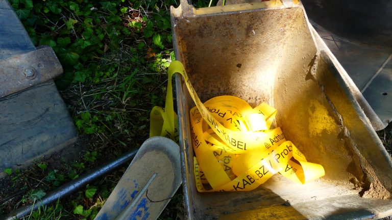 Ein Baggerlöffel enthält gelbes Warnband, auf dem "Achtung Kabel" steht. Neben dem Löffel liegen ein grauer Schutzhelm und andere Werkzeuge im Gras. Im Hintergrund sind grüne Pflanzen zu sehen.