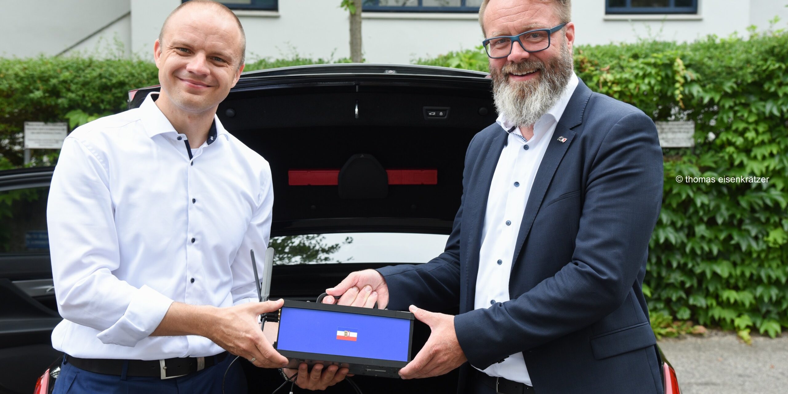 Zwei Männer stehen vor einem geöffneten Kofferraum. Beide tragen Hemden mit langen Ärmeln. Sie halten gemeinsam ein Gerät. Im Hintergrund sind eine Hecke und ein Gebäude mit Fenstern zu sehen.